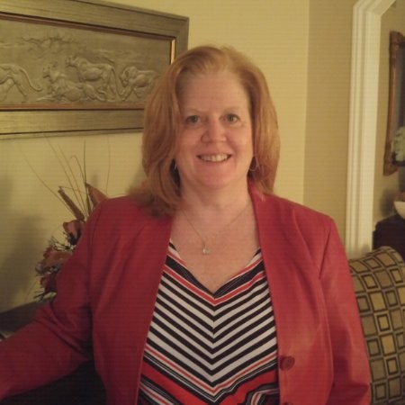 Smiling woman with shoulder-length red hair wearing a red blazer and striped blouse, indoors with decorative wall art behind.